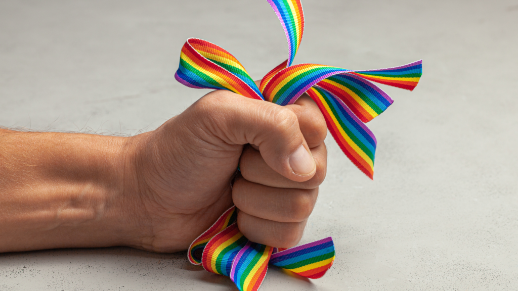 Symboldbild für Queerfeindlichkeit und Homophobie: eine Faust hällt aggressiv fest ein paar Regenbogenbänder