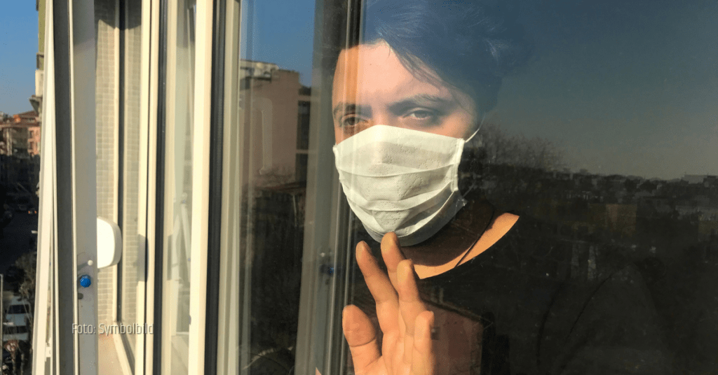Ein Mann steht während der Corona-Pandemie in seiner Wohnung beim Fenster. Er trägt eine Maske und schaut sehnsüchtig nach draußen.