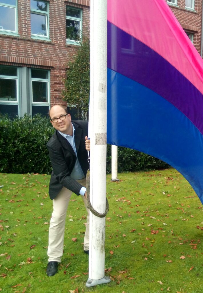 Frank Thies hisst Flagge der bisexuellen Menschen an Fahnenmast auf grüner Wiese.