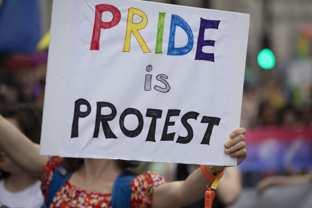 Person hält ein Schild mit der Aufschrift „Pride is Protest“ bei einer Pride-Demonstration hoch.