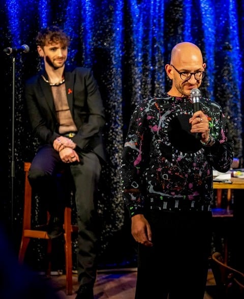 Torsten Poggenpohl (rechts) liest bei einem Charity-Abend auf der Bühne, neben ihm sitzt ein weiterer Künstler, stimmungsvolle Beleuchtung im Hintergrund.