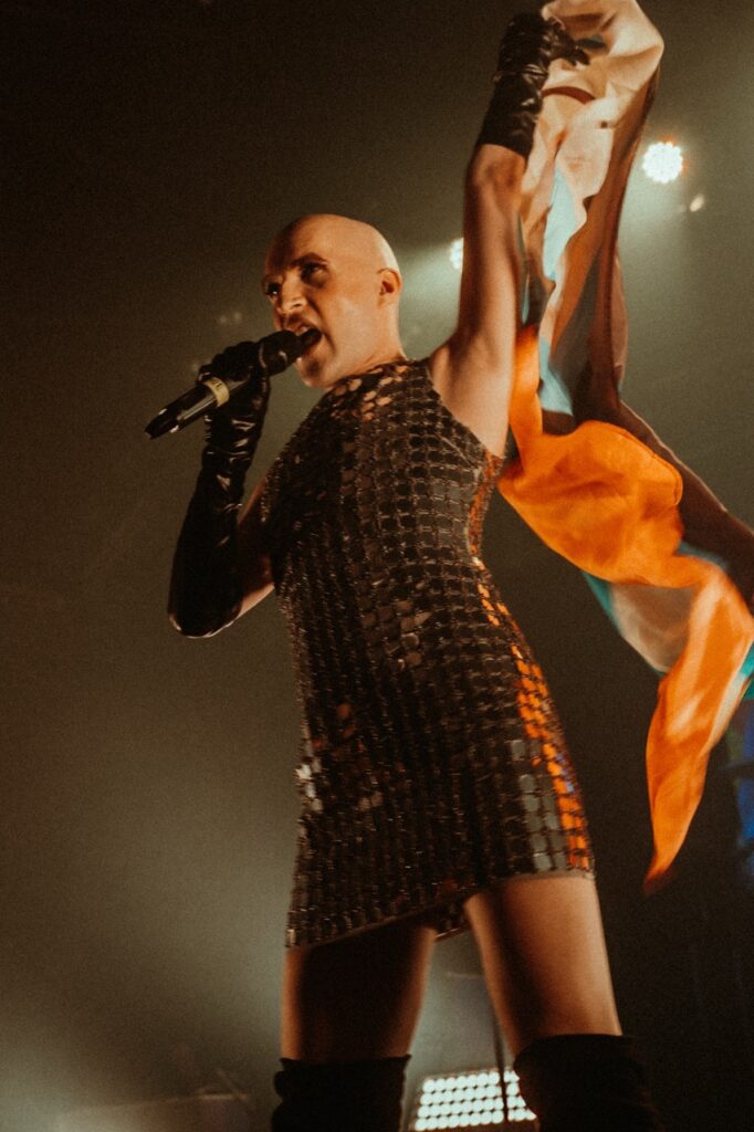 Person in schwarzem Bühnenoutfit singt in ein Mikrofon und hält eine Pride-Flagge hoch.