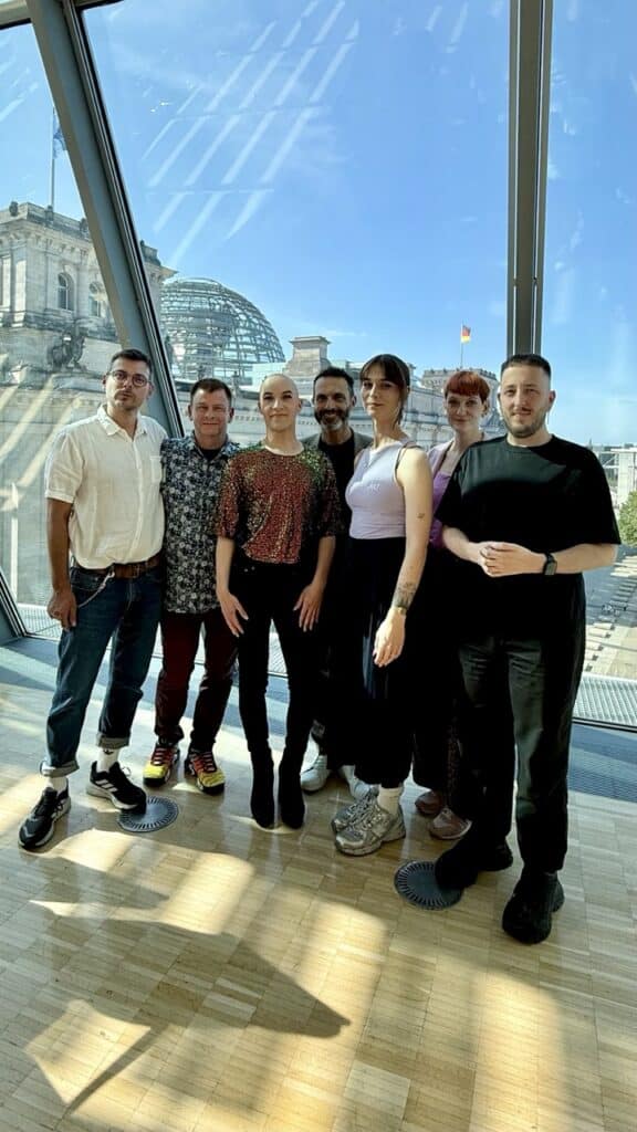 Sieben Personen stehen gemeinsam vor einer Glasfront mit Blick auf die Reichstagskuppel in Berlin.