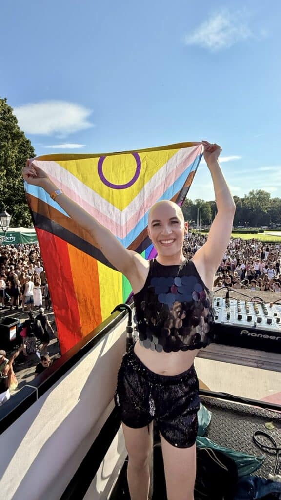 Person hält auf einer Bühne beim CSD eine bunte Pride-Flagge mit intergeschlechtlichem Symbol hoch. Im Hintergrund ist eine große Menschenmenge zu sehen.