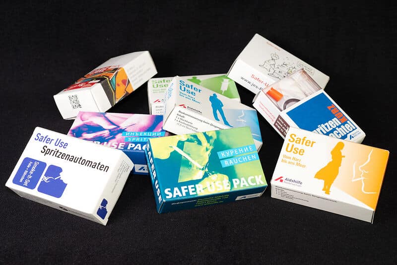 Verschiedene Safer Use Packs und Informationsmaterialien der Deutschen Aidshilfe zum risikoärmeren Drogenkonsum auf schwarzem Hintergrund.