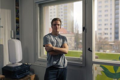 Danilo stands with his arms folded in front of a window in his flat in Berlin and looks into the camera.