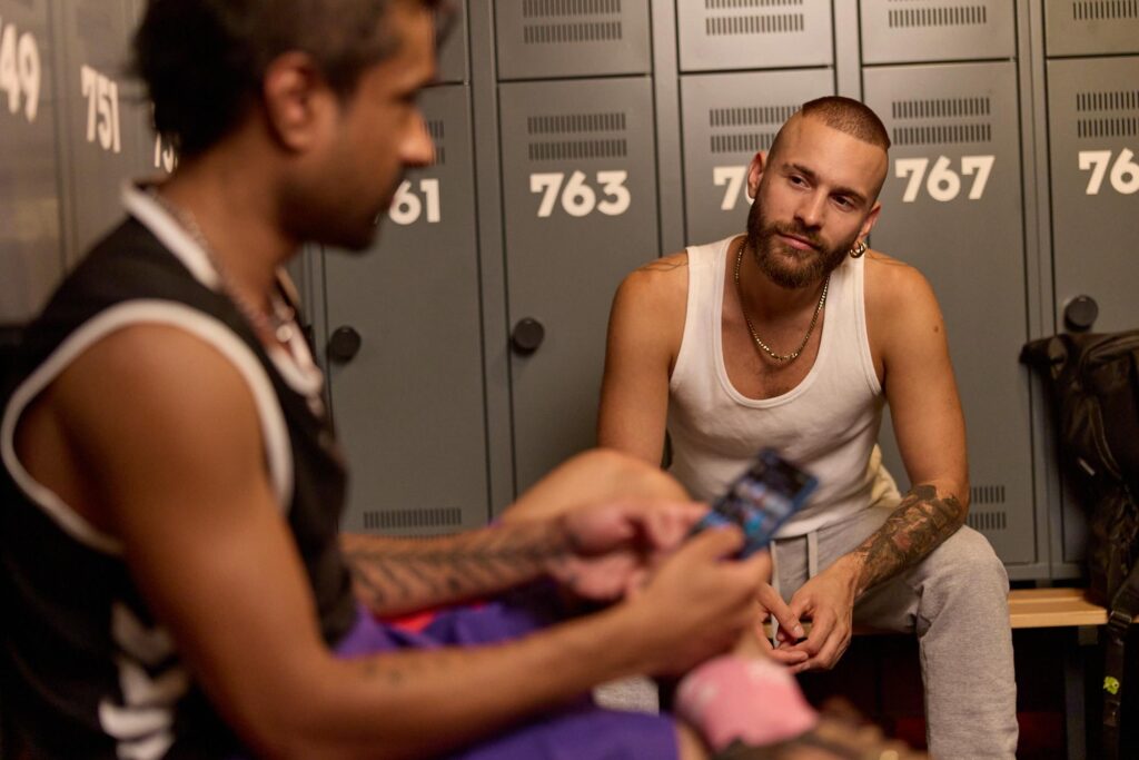 Zwei Männer sitzen in der Umkleide, einer schaut aufs Smartphone – Symbol für Grindr-Chat und Ghosting.