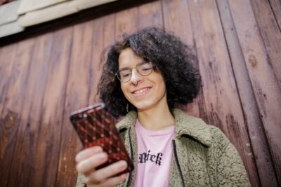 Lizzy stands in front of a wooden façade and looks at her mobile phone with a smile
