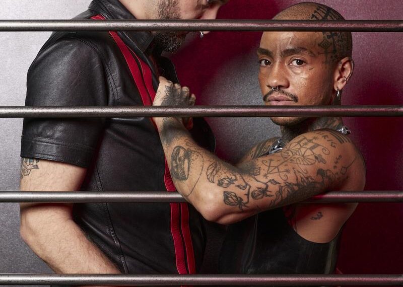 Two people in close physical proximity behind metal bars, one person looking directly into the camera, both wearing black outfits